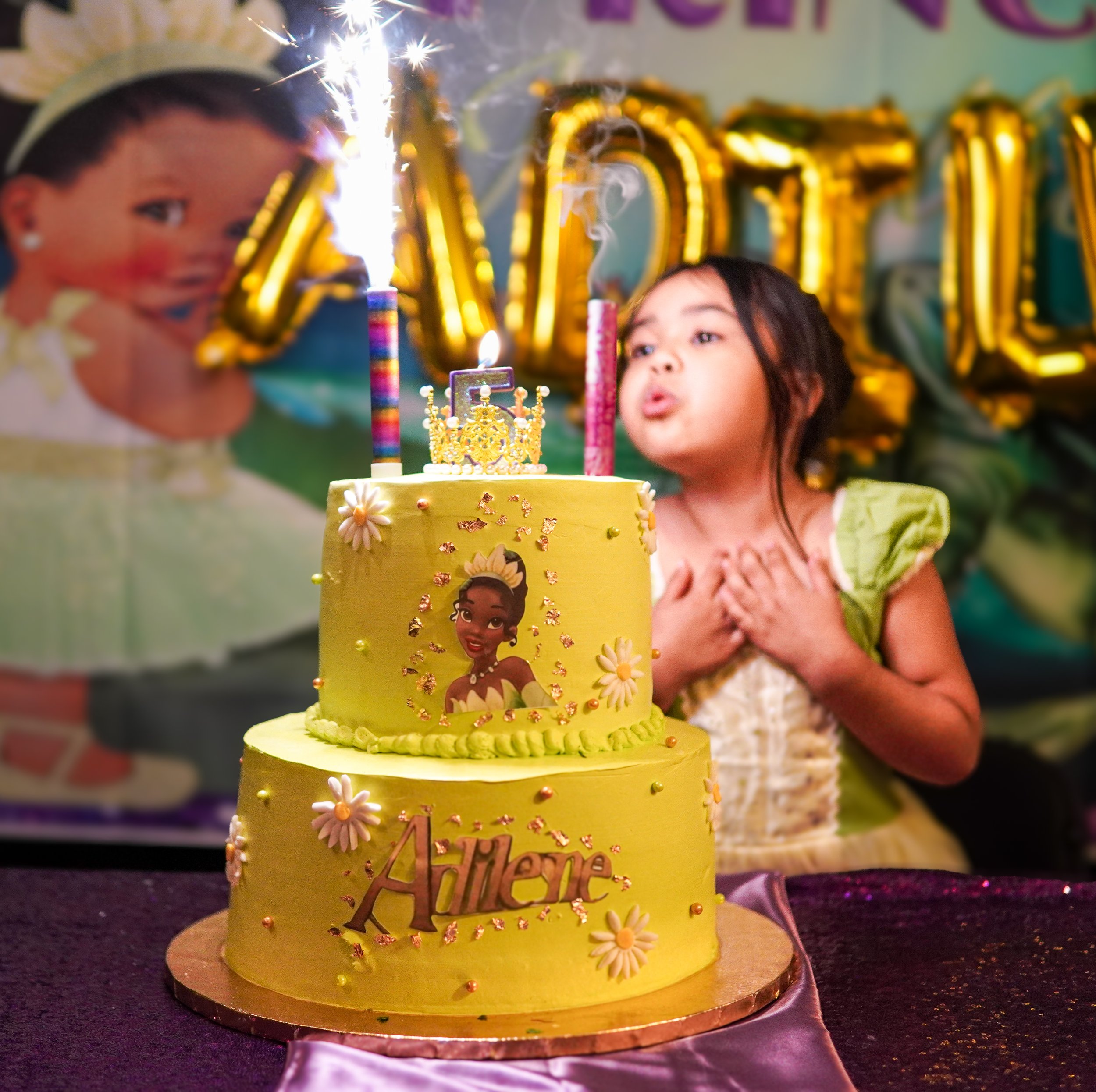 A young girl celebrating her birthday with a yellow birthday cake decorated with princess and flower images, topped with lit candles and sparklers, with gold letter balloons in the background spelling her name, Ahlene.