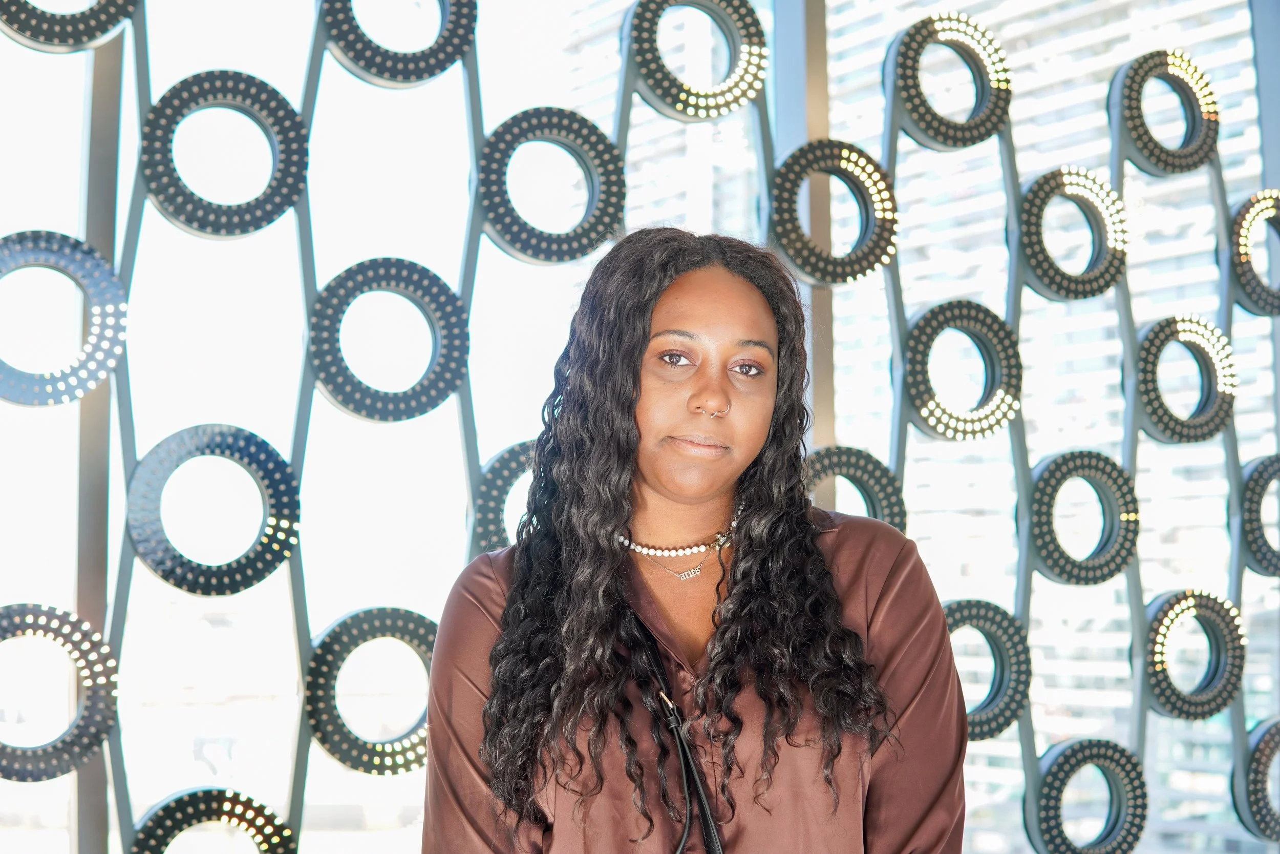 A woman with long curly hair, wearing a brown satin blouse, silver jewelry, and a lanyard, stands in front of a modern art wall with circular light fixtures, in a bright room with large windows.