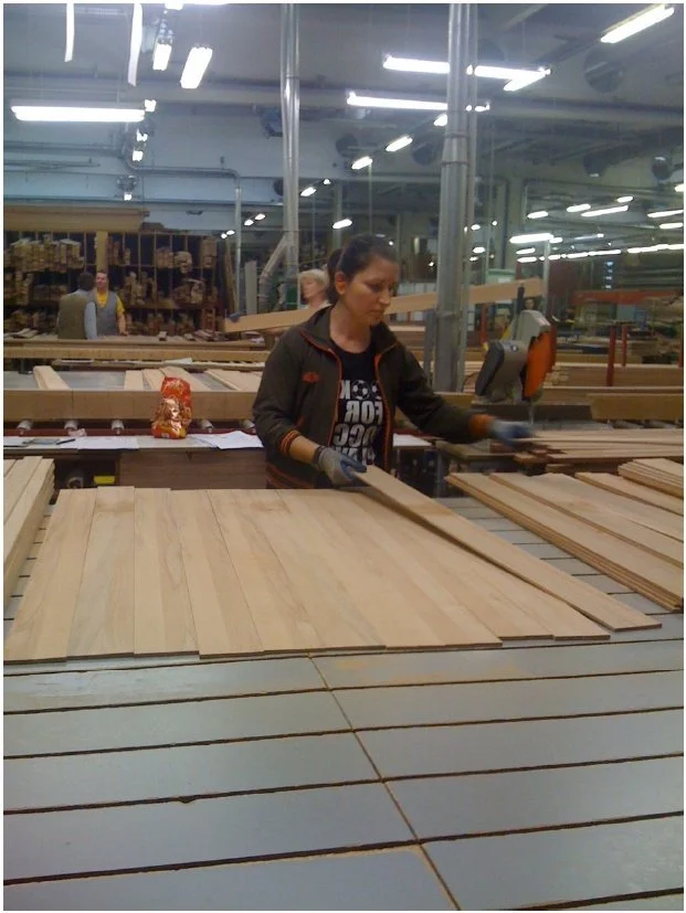 A woman working in a woodworking factory, cutting or preparing wooden planks. There are several stacks of wood around her and others working in the background.