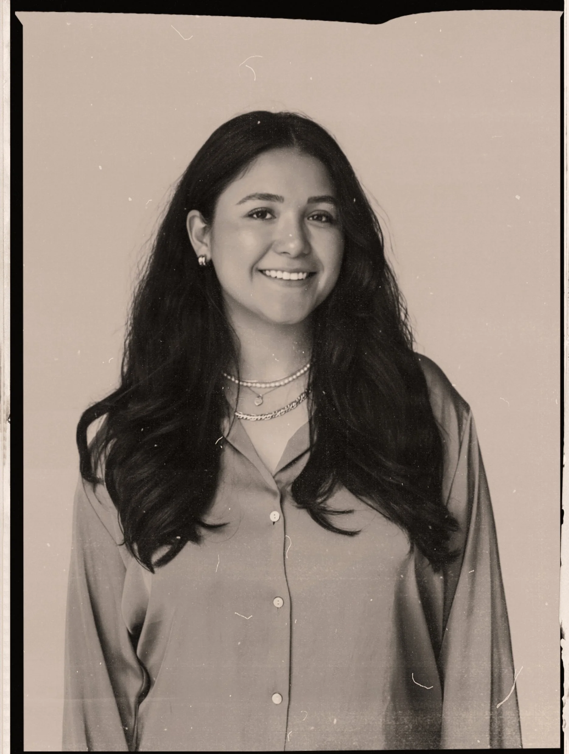 Black and white portrait of a young woman with long, wavy hair smiling, wearing earrings, layered necklaces, and a button-up shirt.