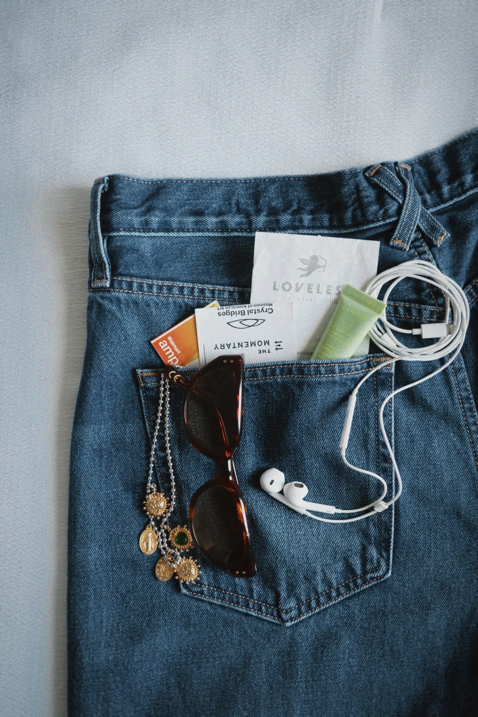 A pair of blue jeans with items in the back pocket: earrings, sunglasses, a business card, a flyer, a small tube of lotion, and white earphones.