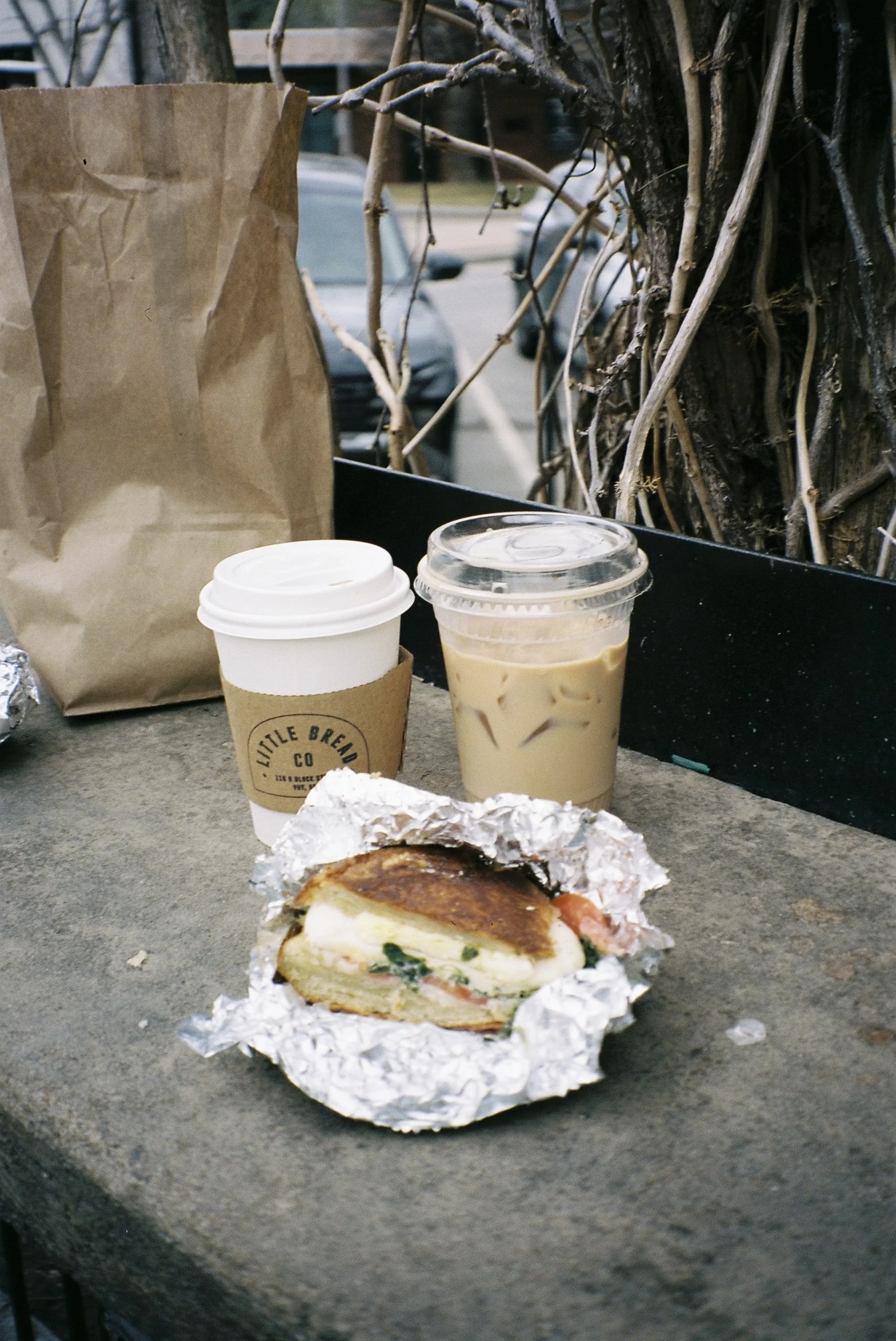 An outdoor scene with a sandwich wrapped in foil, a white to-go coffee cup with a cardboard sleeve, a clear plastic cup with iced coffee, and a brown paper bag on a concrete surface, with a tree and parked cars in the background.