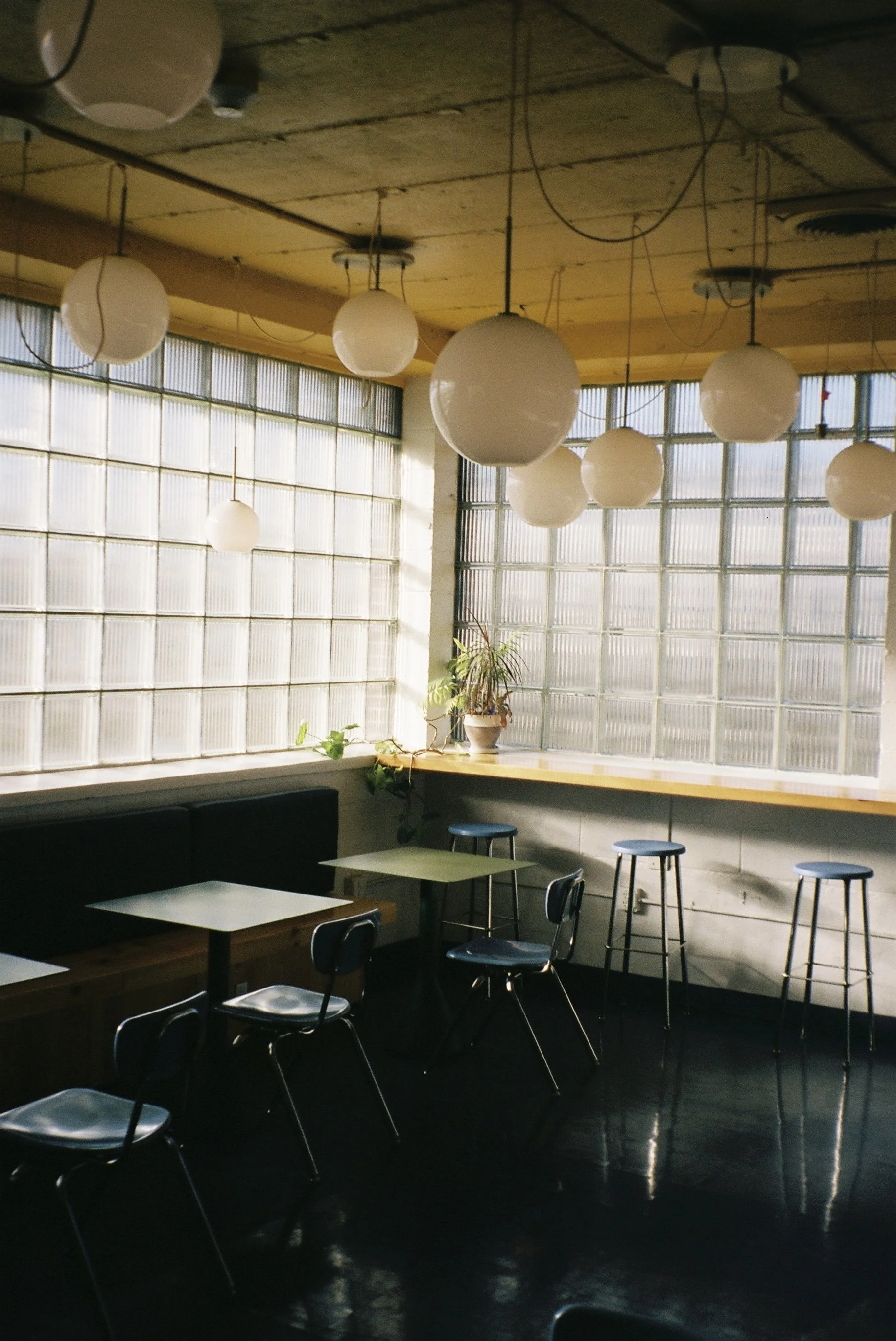 An empty indoor cafe with large glass block windows, hanging spherical pendant lights, a long wooden counter with plants, and a few tables and chairs.