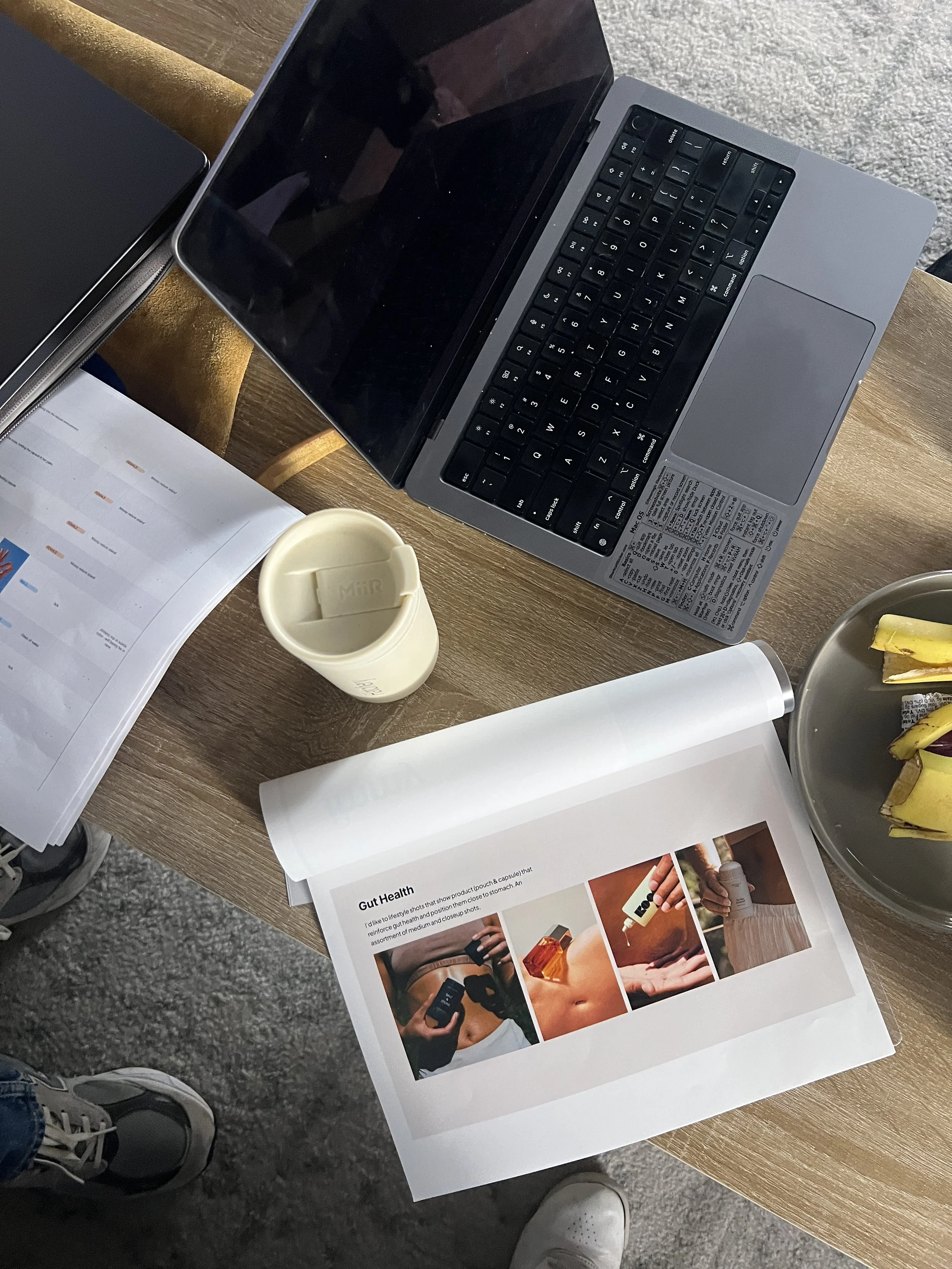 A wooden table with an open laptop, a printed magazine page about gut health, a white cup, a plate with banana peels, and some books or notebooks.