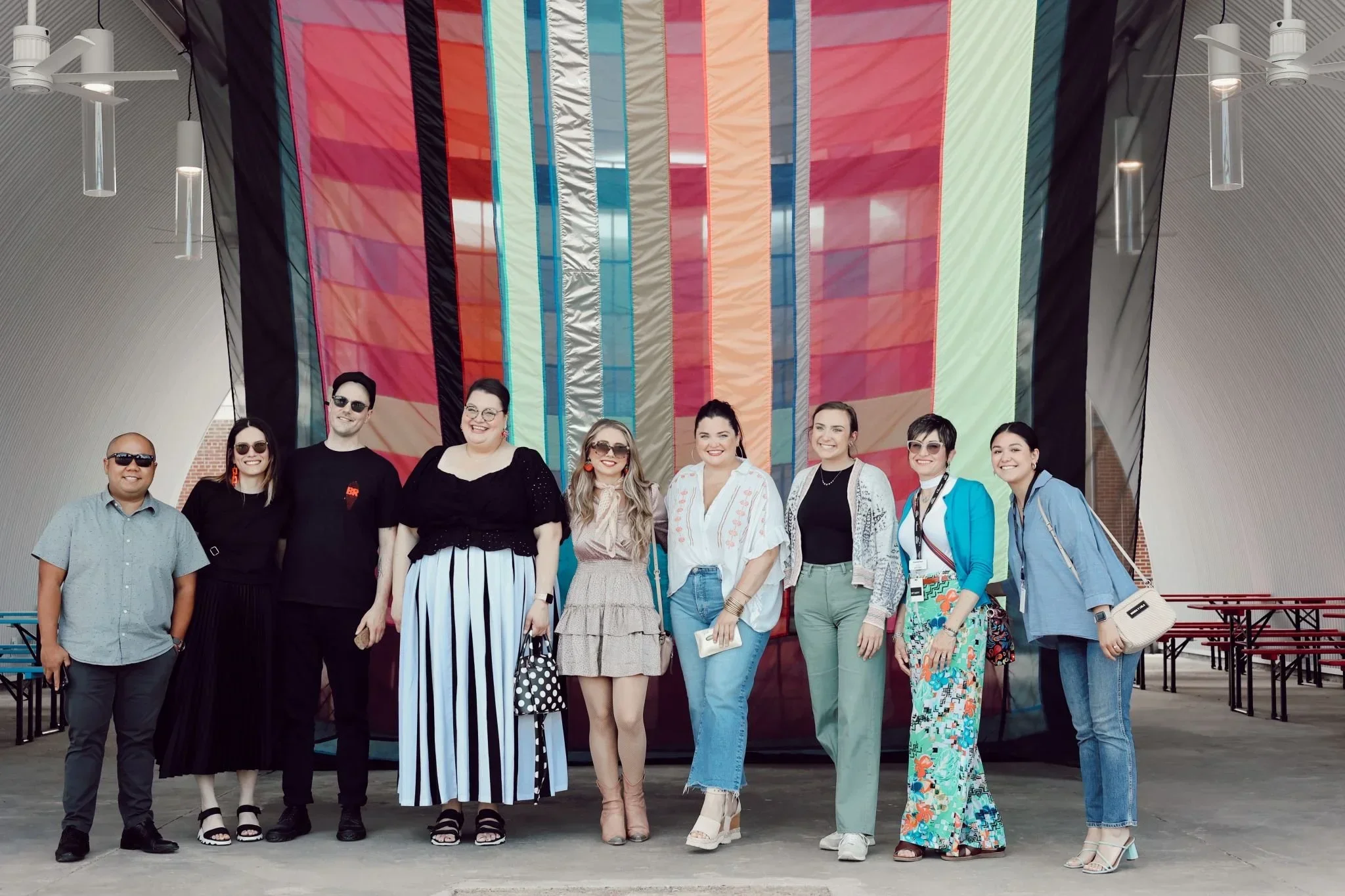 A group of ten diverse people standing in a line outdoors in front of colorful hanging fabric or banners, smiling at the camera.