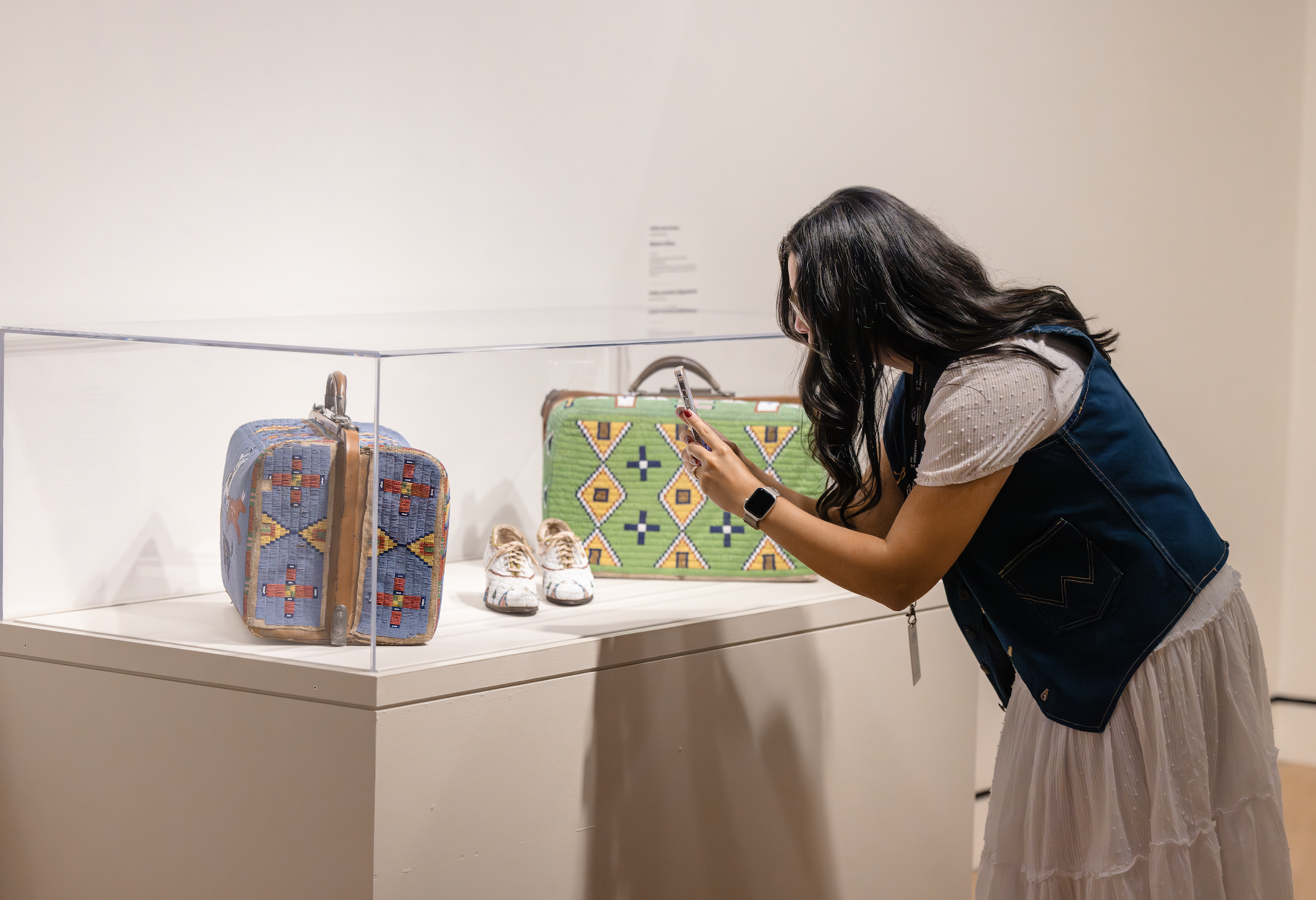 A woman with long dark hair wearing a beige dress and denim vest looking at exhibit items in a glass case in an art gallery.