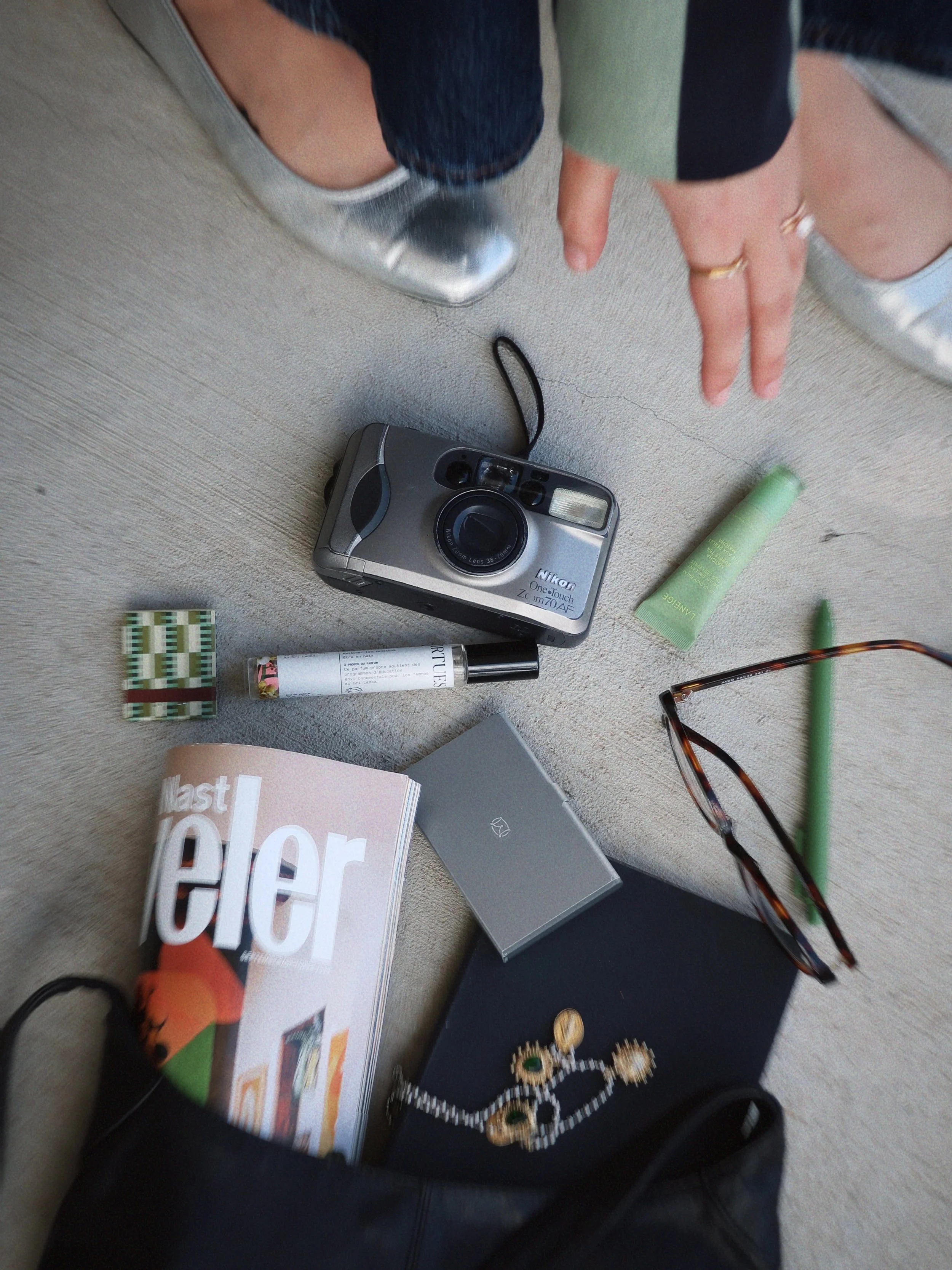Collection of personal items scattered on a beige carpet including a silver camera, a pair of eyeglasses, a magazine, a small green tube, a tube of lotion, a lipstick, a small jewelry box, a black pouch, and jewelry pieces.