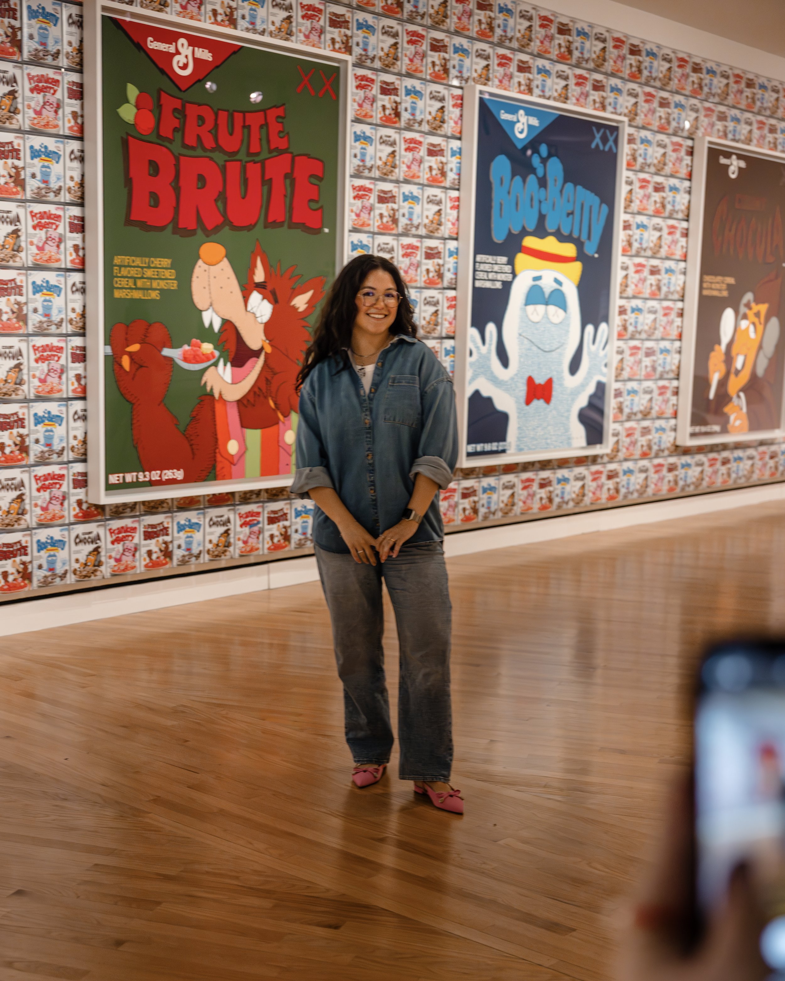 A woman standing in an art gallery with wallpaper made of boxes of cereal and three large colorful posters behind her, showcasing cereal characters and branding.