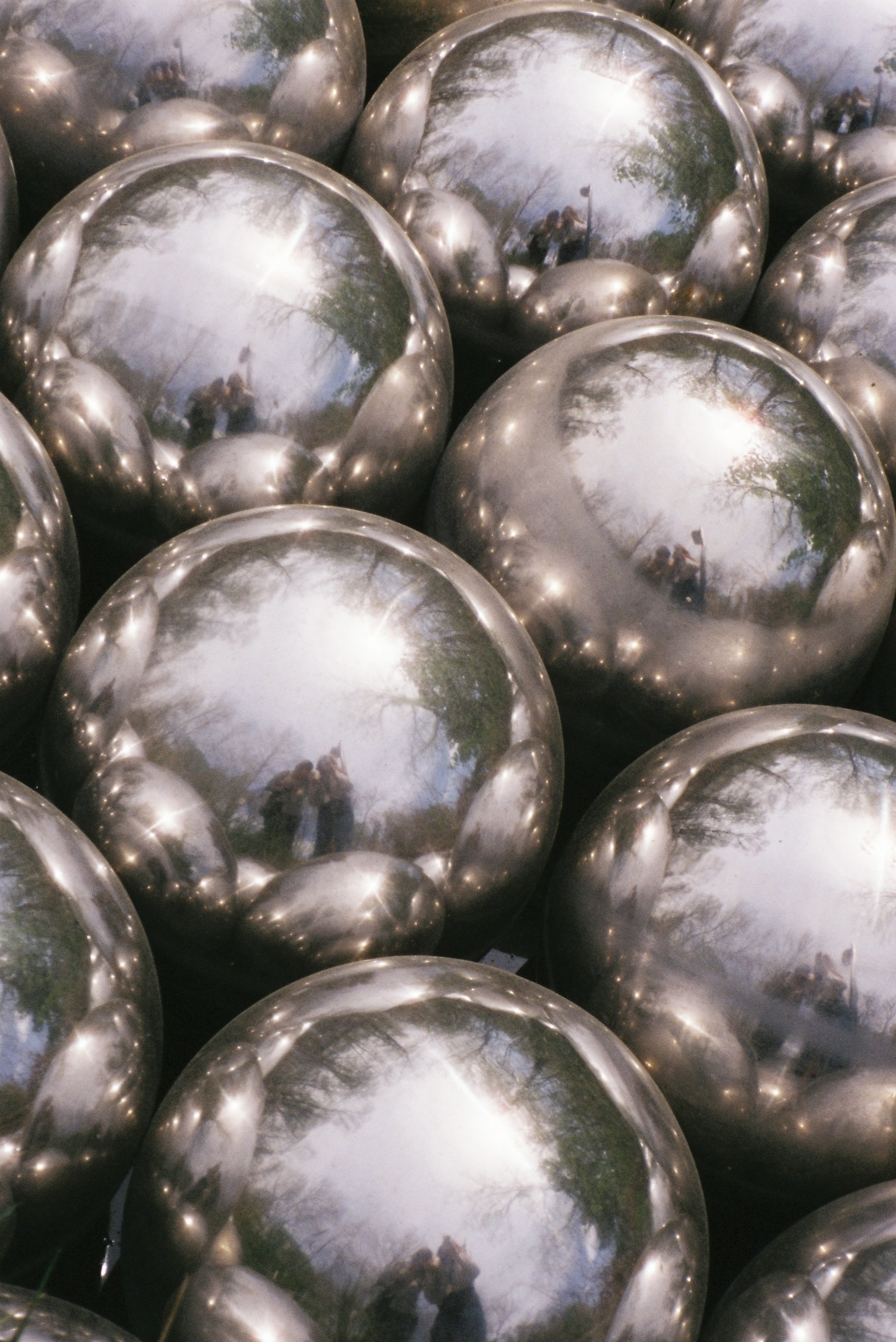 Numerous shiny metallic spheres reflecting the surrounding trees and sky, with some people and a flag visible in the reflections.