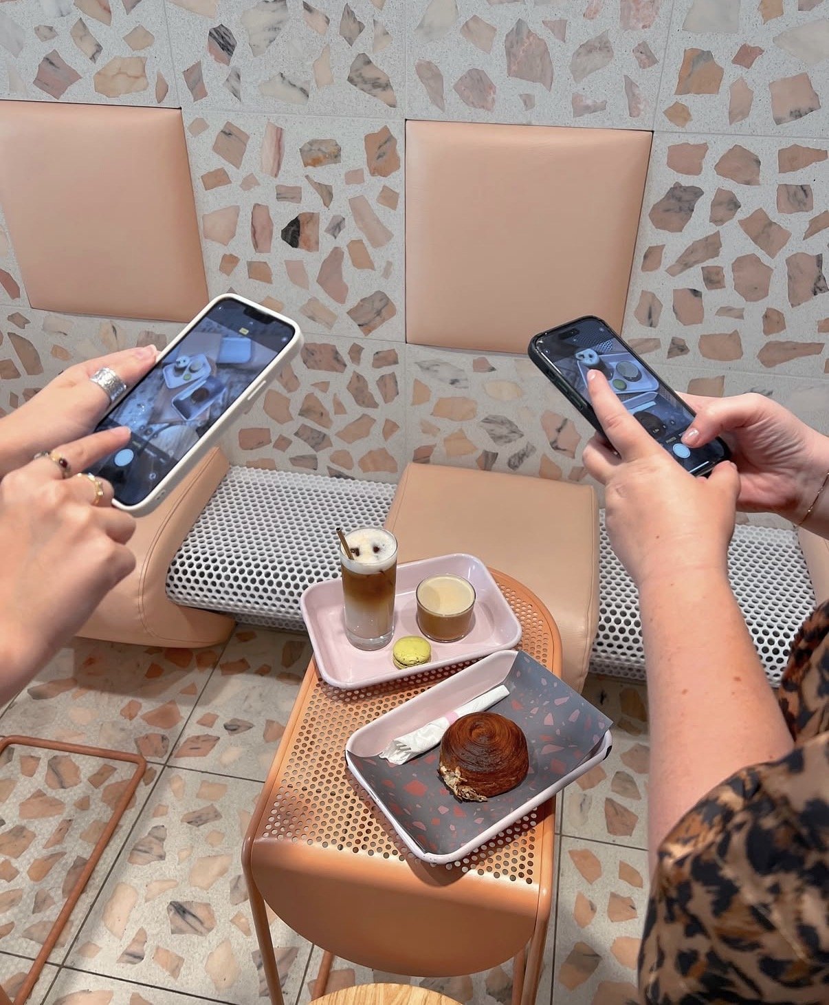 Two people taking photos of a table with pastries and beverages in a cafe with terrazzo walls and peach-colored seating.