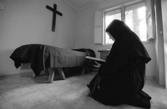 A woman dressed as a nun kneeling and reading a book in a room with a bed and a crucifix on the wall.