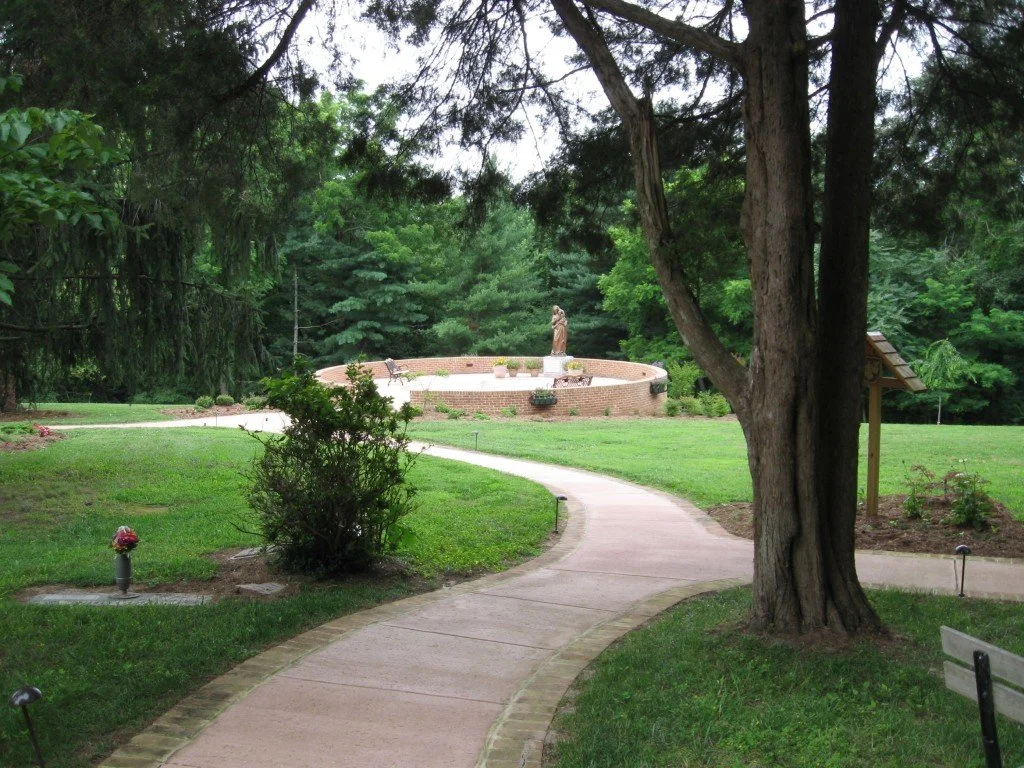 A curving concrete pathway through a park with lush green grass and trees, leading to a circular brick fountain with a statue on top, surrounded by trees and shrubbery.