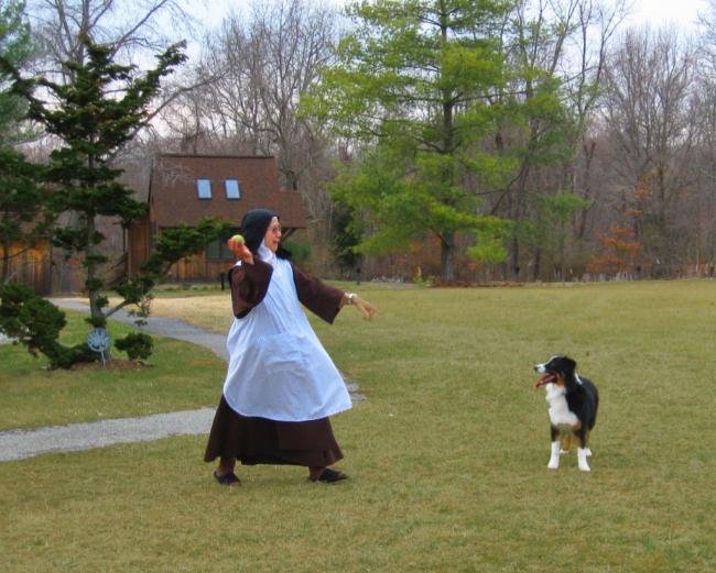 A person dressed as a nun holding a ball, standing on a grassy lawn near a black and white dog, with trees and a house in the background.