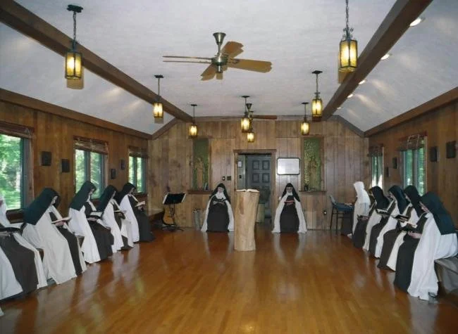 Group of nuns dressed in black and white habits seated in pews, facing each other, inside a wooden chapel or meeting room with large windows, hanging lamps, and a wooden polished floor.