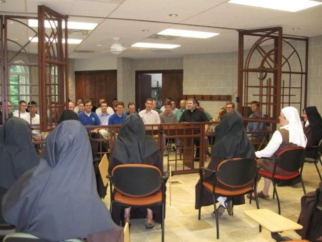 A group of women dressed in black and white hijabs seated in a circle in a room with a mixed-gender audience listening to a discussion or presentation.