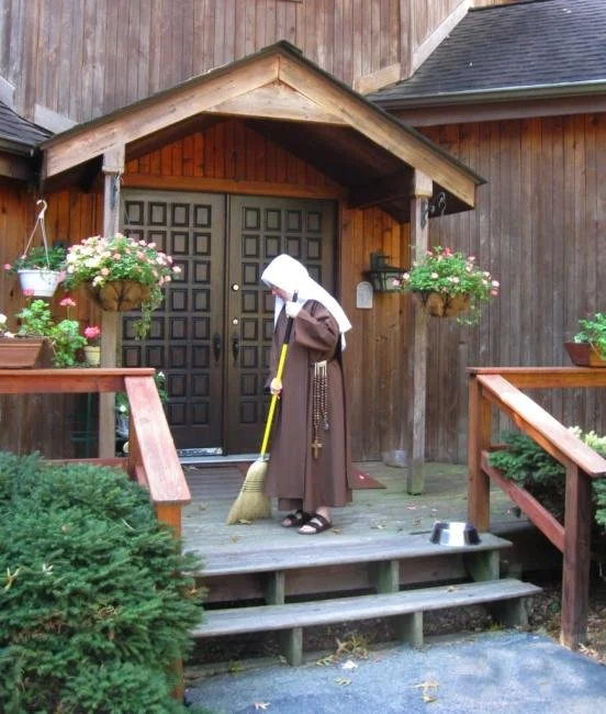 A person in a brown dress, white headscarf, and sandals sweeping the wooden porch in front of a house with dark double doors, surrounded by hanging flower baskets and plants.