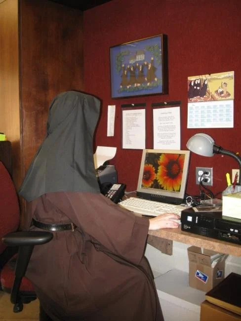 A person wearing a head covering sitting at a desk with a computer, phone, and desk lamp in a room with red walls and framed pictures.