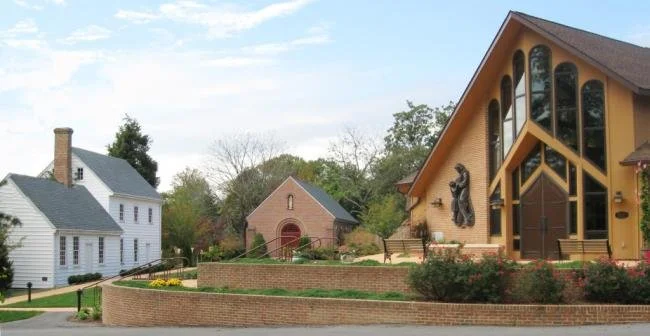 A church with large stained glass windows and a wooden sculpture outside, surrounded by greenery and a brick wall.