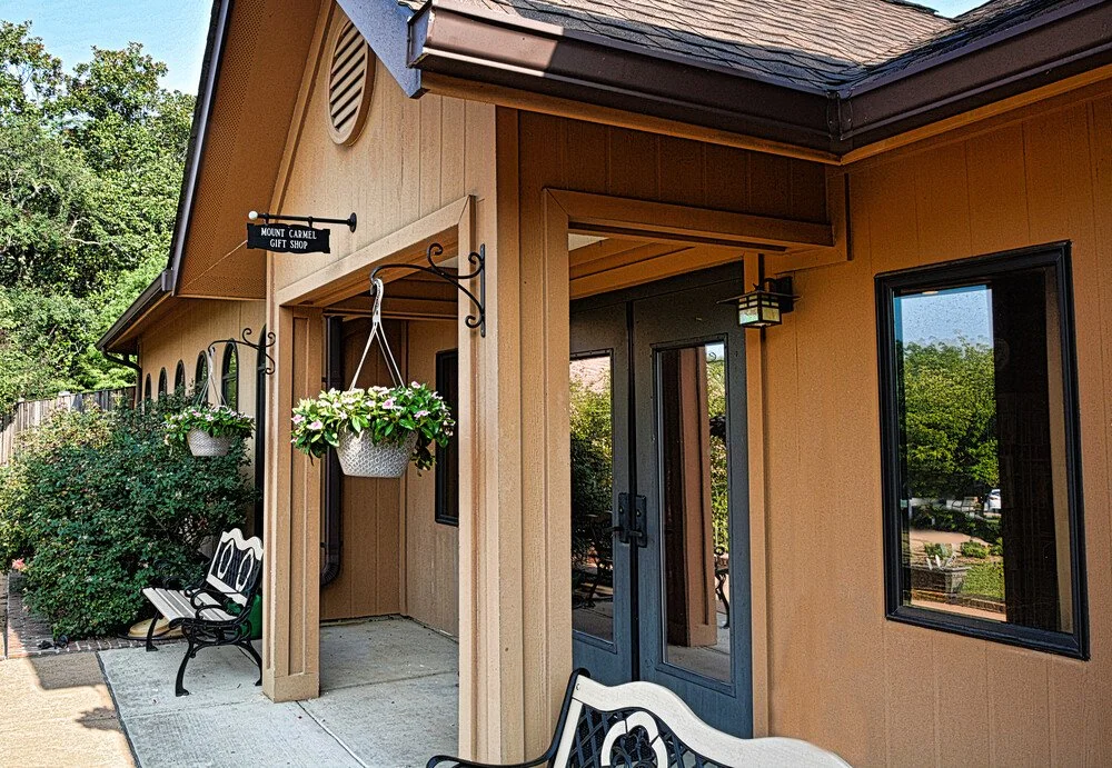 The exterior of a tan building with a sign that reads 'Mount Carmel Gift Shop,' a hanging flower basket, a bench with another flower basket, and a glass door with surrounding windows.