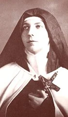 Black and white photo of a woman with long dark hair, wearing a dark cloak and a light-colored garment underneath, holding a crucifix cross.