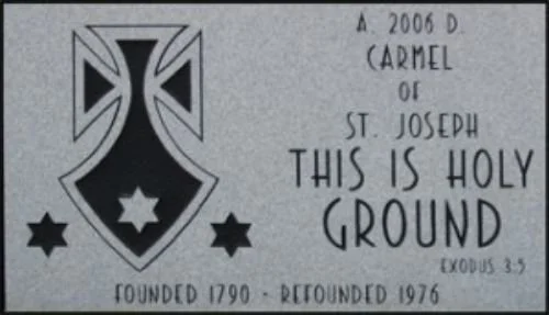 A black and white sign featuring a military-style insignia with a cross and stars, text about Carmel of St. Joseph, and references to biblical scripture and church history.