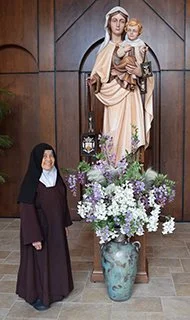 A nun standing with a large floral arrangement in front of a religious statue of a woman holding a child, in a church setting.