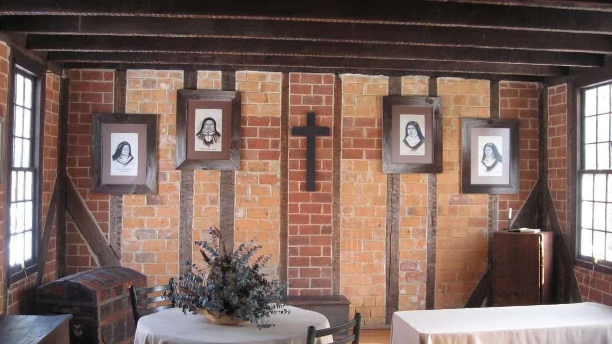 A rustic brick room with four framed religious portraits of nuns on the wall, a wooden cross in the center, a table with a floral arrangement, and windows on each side.