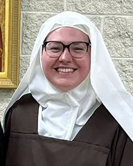 A woman dressed as a nun, smiling, wearing glasses, in front of a beige brick wall with a yellow window frame.