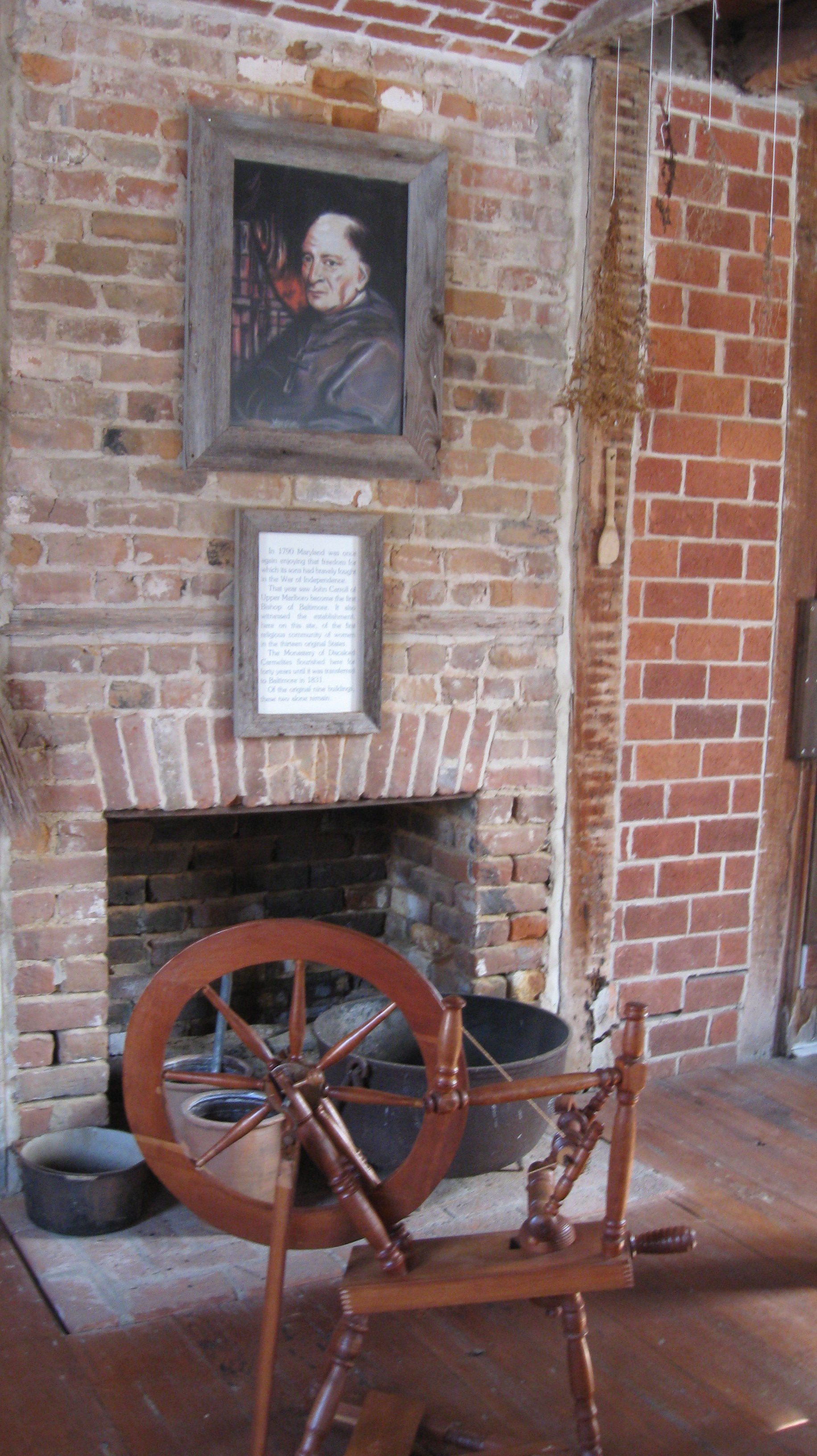 A room with a brick fireplace, a spinning wheel, and framed artwork on the wall. The artwork is a portrait of a man with white hair in a leather jacket. There is an informational plaque below the artwork, and the room has brick walls and wooden flooring.