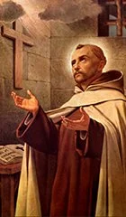 A priest standing with hands raised in prayer inside a church, with a large cross behind him.