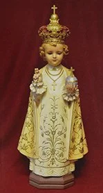 Statue of a young girl dressed as a queen, wearing a gold crown and ornate robes, against a red background.