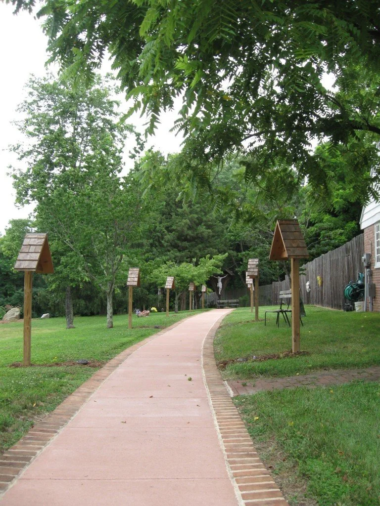 A winding sidewalk in a green backyard with trees, wooden birdhouses on posts, a bench, and a wooden fence.