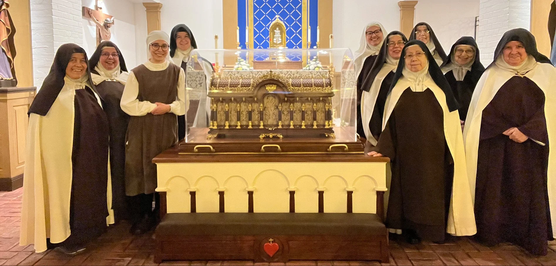 A group of nuns standing around a religious relic inside a church. They are wearing black and white habits. The relic is displayed in a glass case on a table decorated with gold accents.