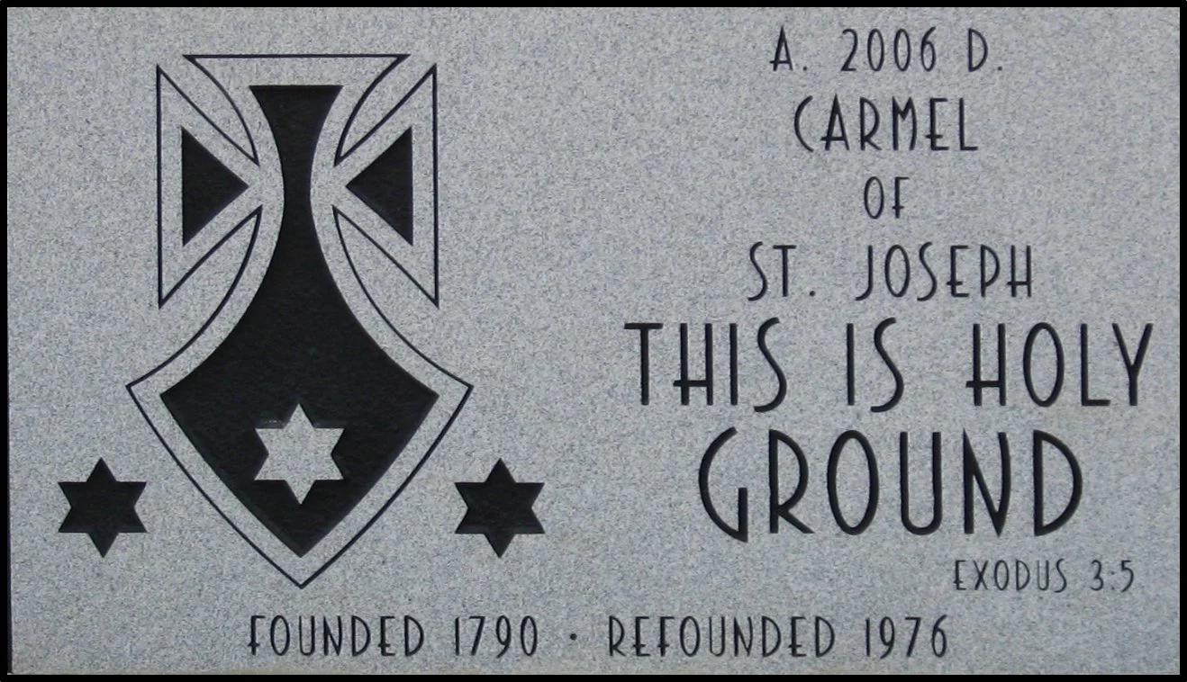 A plaque with a stylized black and white emblem and text that reads: "A. 2006 D. CARMEL OF ST. JOSEPH THIS IS HOLY GROUND EXODUS 3:5. FOUNDED 1790. REFOUNDED 1976."
