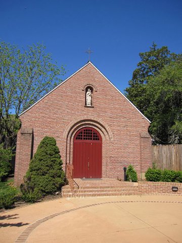 A small brick church with a red door, arched window, and a cross on top, surrounded by green trees and a dirt pathway.