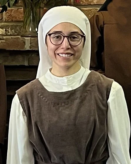 A woman dressed as a nun with glasses, a white head covering, and a brown habit, smiling in front of a brick fireplace.