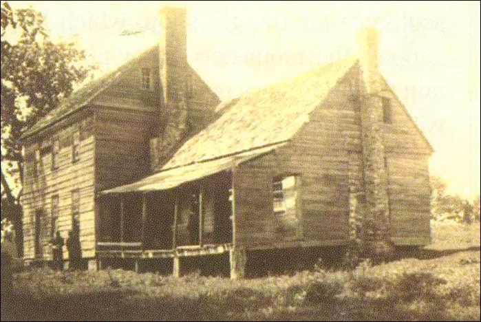 Old wooden house with a porch, chimney, and damaged roof, possibly a historic or abandoned building.