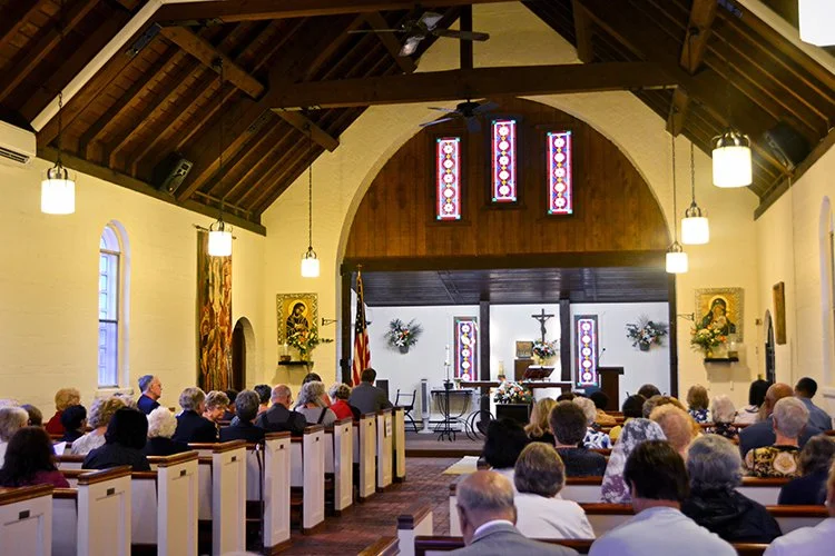 A church interior with a congregation seated on white pews facing the altar. The church has high wooden ceilings, hanging lights, stained glass windows, and religious artwork on the walls. The altar area features a cross, flowers, and religious items.