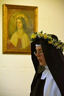 A woman dressed as a nun with a black veil and white habit, wearing a flower crown, standing in front of a yellow wall with a framed painting of Jesus Christ in the background.
