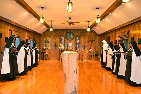 A group of nuns in black and white habits standing in a semi-circle inside a wooden chapel, reading from books.