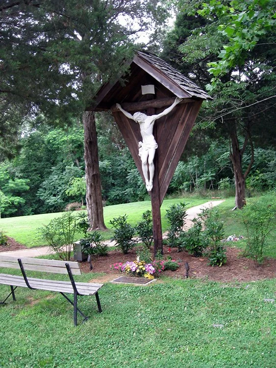 Crucifix sculpture of Jesus Christ in a wooden shelter in a park with trees, benches, and flower beds.