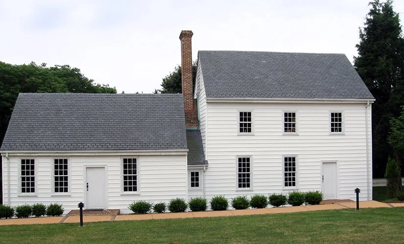 White house with gray shingled roof, multiple windows, and a chimney, surrounded by green grass and bushes.