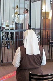 A woman with a white cloth over her head sitting at a table in a restaurant, watching through large glass windows at a man and woman near a bicycle outside.