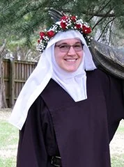 Woman wearing a black graduation gown with a white headscarf and a floral wreath on her head, standing outdoors in front of a wooden fence and trees.