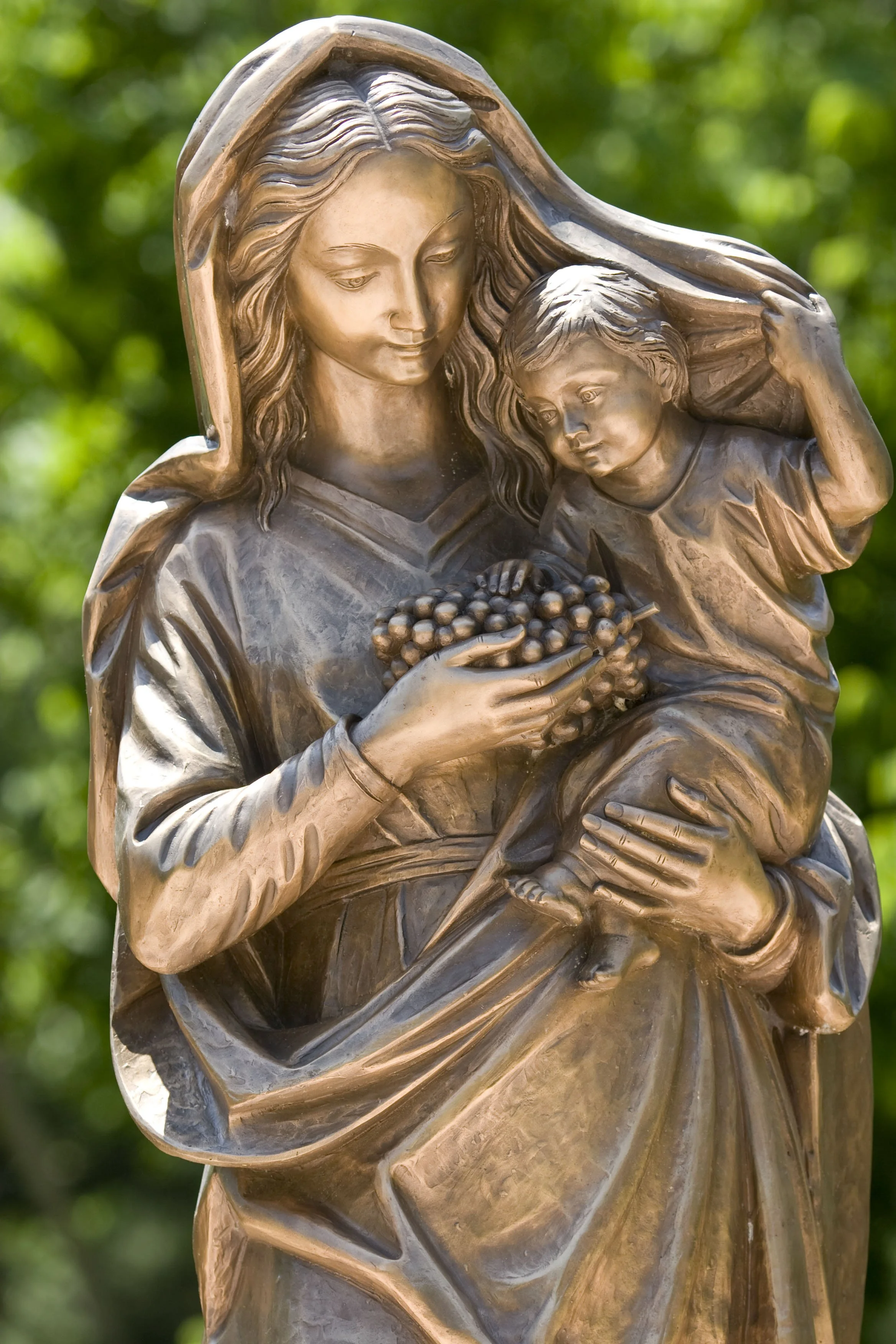 Close-up of a bronze sculpture of the Virgin Mary holding baby Jesus, with a green leafy background.