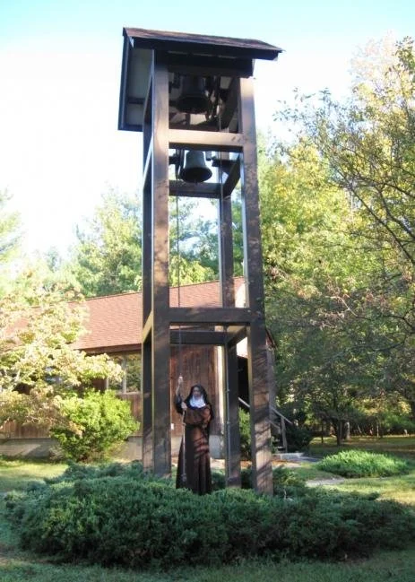 A person standing underneath a tall wooden bell tower with a bell inside, surrounded by trees and a house in the background.