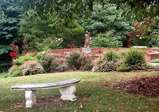 A garden scene with a stone bench in the foreground, surrounded by bushes and trees, with a brick wall and flowering plants in the background.