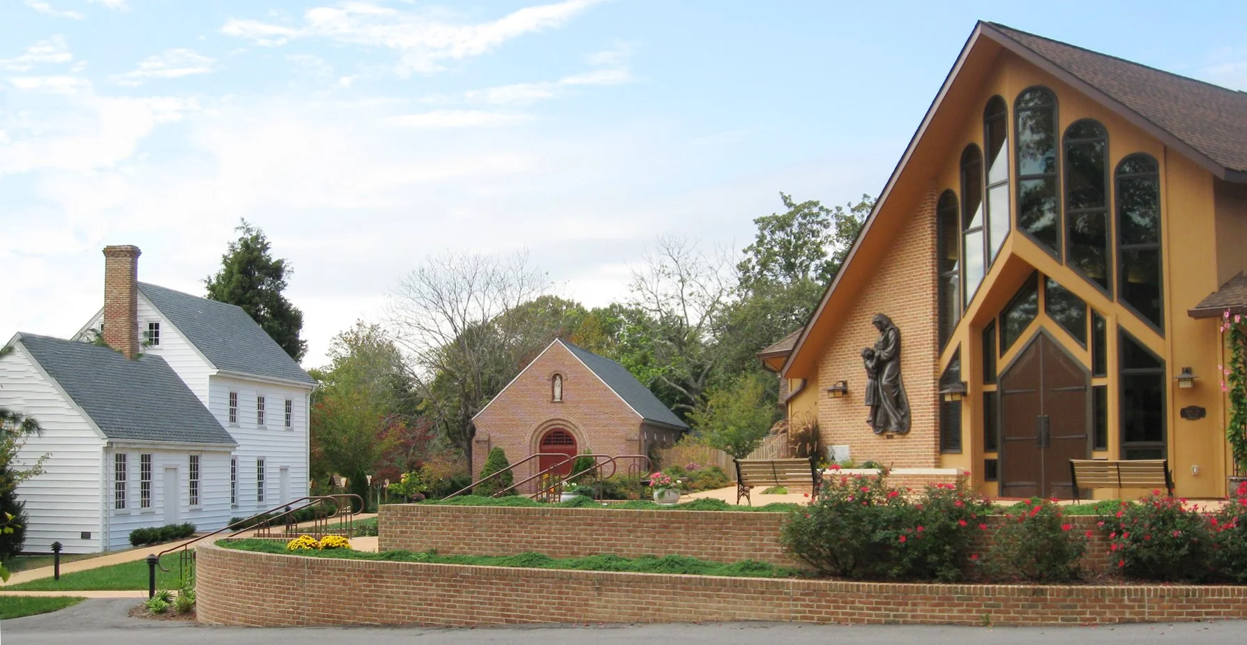 A church with a brick facade and large, arched windows, and a white house with grey roof and chimney in a landscaped area with benches and flowering bushes.