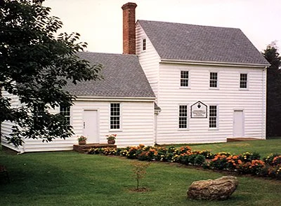 White two-story house with a gray roof, surrounded by a neatly maintained lawn and colorful flower beds.