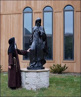 A woman in a hijab and long dress standing outside near a statue of a religious figure, holding hands with the statue.